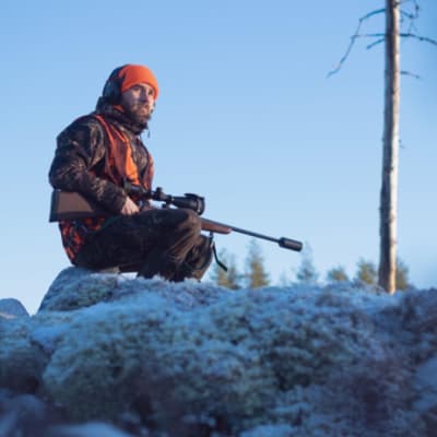 Istuva metsästäjä huurteisessa maisemassa, jossa näkyy lisäksi taivasta vasten kukkula ja yksi ohuehko puunrunko.