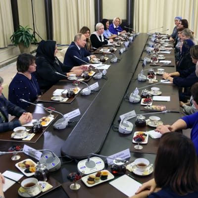 Vladimir Putin sitter i ett bord och talar med flera kvinnor. De har neutrala ansiktsuttryck.