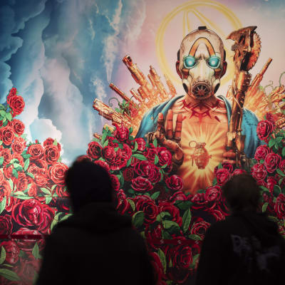 Människor går förbi en stor reklam för Bordelands spelet. På reklamen ses en person omringad av vackra röda rosor och vapen. Han har på sig en mask där ögonen lyser blåa.