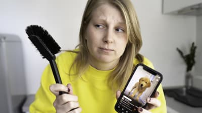 Huolestuneen näköinen nainen katsoo tiskiharjaa ja pitää kädessään kännykkää, jonka ruudulla on koiravideo.