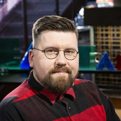 Mies punaruudullisessa paidassa ja silmälaseissa lähikuvassa katsoo kameraan, taustalla Yle Perjantain värikkäitä lavasteita.
