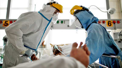Två vårdare i skyddsutrustning står vid en sjukhussäng där en coronapatient ligger. Patienten har en andningsmask på.