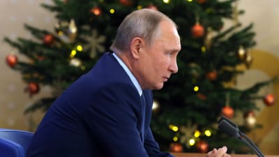 Vladimir Putin sitter vid sitt bord i sidoprofil och har en dekorerad julgran i bakgrunde. Putin har knäppt sina händer.