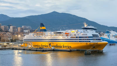 Ett gulvitt kryssningsfartyg ligger i en hamn i ett medelhavslandskap. På den gula delen står det med mörka bokstäver Corsica Ferries - Sardinia Ferries.