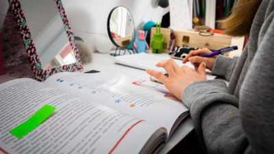 En ung flicka studerar på distans. Hon har framför sig en bok på ett skrivbord.