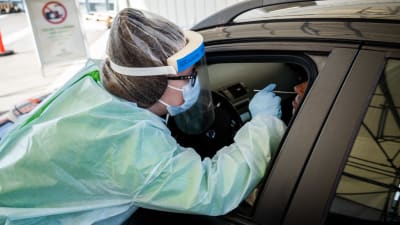 En vårdare i skyddsutrustning tar ett coronatest på en person som sitter i en bil.