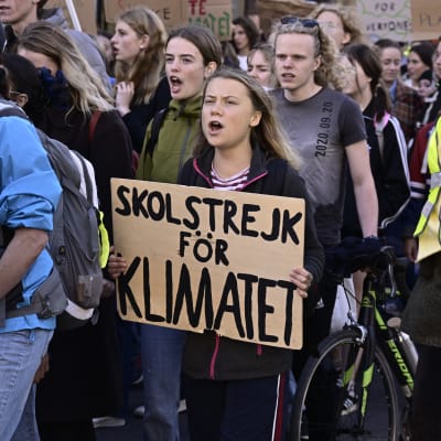 Greta Thunberg går bland demonstranter. Hon håller en skylt där det står skolstrejkt för klimatet.