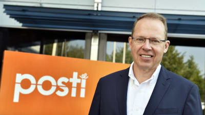 Heikki Malinen, en äldre man i blå kostym med glasögon. Han står framför en orange skylt med texten Posti.