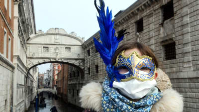 Ett barn iklätt ansiktsmask med en stor blå fjäder och andningsskydd vid en av Venedigs broar över kanalen.
