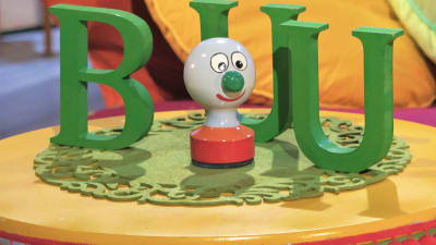 BUU-klubbens maskot BUU-stämpeln står på ett gult bord. Bakom stämpeln finns gröna bokstäver av trä B U och U. BUU-stämpeln är en figur med grön näsa, glad röd mun, stora ögon och en röd fot.