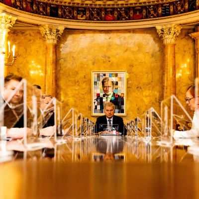 Sauli Niinistö sitter vid ett bord med regeringen i en guldsal. Bakom honom hänger ett porträtt av honom.