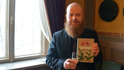 En man står inomhus vid ett fönster. Han ler och tittar mot kameran. I ena handen håller han ett glas med skumvin, i den andra en bok med namnet Sömnö.