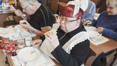 Pensionärer sitter vid pulpeter och äter gammaldags skollunch bestående av välling och knäckebröd.