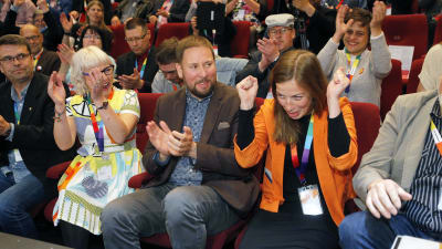 Li Andersson i orange jacka jublar med händerna i knytnävar. Paavi Arhinmäki sitter bredvid henne och applåderar tillsammans med andra partimedlemmar i bakgrunden.
