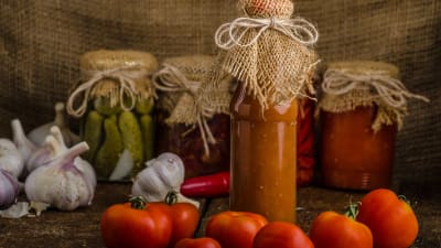 Hemgjord tomatsås i glasburk. I bakgrunden andra inlagda grönsaker och färska tomater och vitlök.