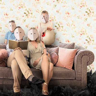 Perhe istuu sohvalla kukkatapetoidun seinän edessä, kaikkien kasvoja peittävät valkeat pahviset naamarit.