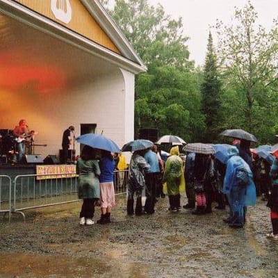 Hämeenlinnan Kaupunginpuiston esiintymislavalla Ultramariini Ämyrockissa vuonna 2004, yleisöä sateenvarjoineen kuuntelemassa.