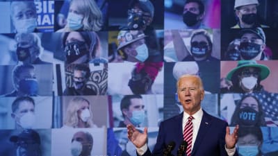 Joe Biden vid mikrofoner, framför en bildvägg med angängare iklädda coronaskydd.