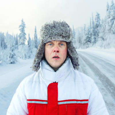 En man i pälsmössa står ensam mitt på en snöig landsväg.
