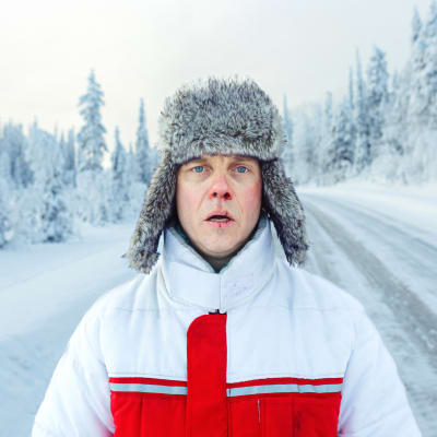 En man i pälsmössa står ensam mitt på en snöig landsväg.