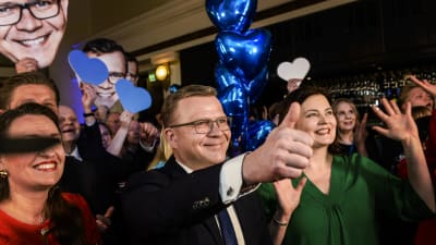 Petteri Orpo omringad av glada människor. Orpo håller upp tummen.