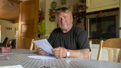 En man i 60-årsåldern sitter vid ett matbord och håller i några dokument. Han tittar in i kameran.