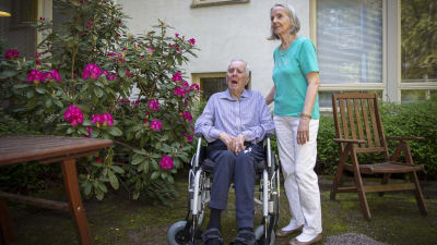 Irmeli och Heikki Laajanen utanför ett vårdhem. Heikki sitter i rullstol och Irmeli tittar på honom och håller handen på hans rygg. Bakom dem finns en blommande buske och utomhusmöbler.