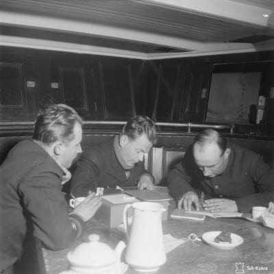 Sähkösanoma muutetaan salakirjoitukseksi. Kolme miestä pöydän äärellä. Paikka: Itämeri. Laivassa?