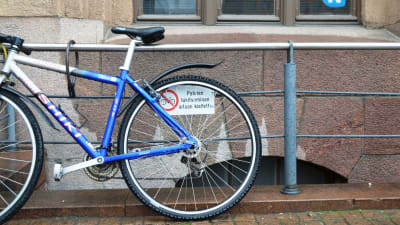 Cykel står parkerad vid ett staket precis vid skylt som visar att det är förbjudet att låsa sin cykel vid staketet.