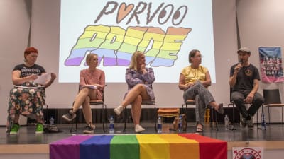 Fyra kvinnor sitter och lyssnar på en man som pratar i en mikrofon. Alla sitter på en scen. Bakom dem står det Porvoo Pride.