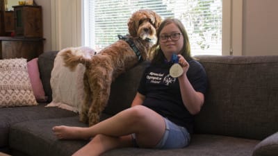 På bilden sitter Alina Skog med en logo-skjorta på sig och håller sin silvermedalj i handen. Bredvid henne på soffan sitter deras hund. 