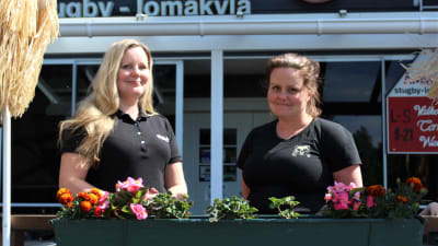 Två leende kvinnor står framför en blomplantering på en terrass.