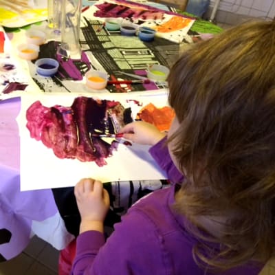 Lapsi piirtää vesiväreillä päiväkodin pöydän ääressä. Lapsella on violetti paita ja keskiruskeat hiukset.