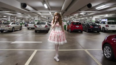En kvinna klädd i så kallade Lolitakläder - en fluffig vit och rosa klänning med spetsar på. Hon har vita högklackade skor och blommor i håret. Hon står i en parkeringsgrotta.