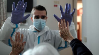 Fysioterapeut i blå plasthandskar och äldre patient sträcker upp sina händer till möte, på äldreboende i Spanien