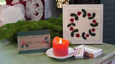Ett grönt slitet träbord som är julpyntat. På bordet finns granris, ett handgjort julkort, ett rött blockljus som brinner, tändsticksaskar med korsstygnsbroderier och en tuftad tavla med en lingonkrans.