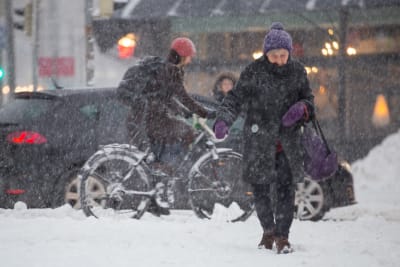 En äldre dam går över vägen i snöoväder, i bakgrunden en cyklist.