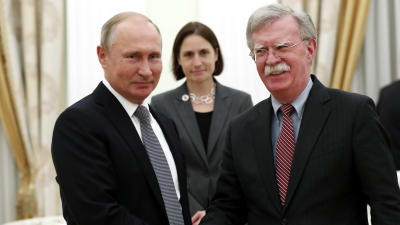 Vladimir Putin och John Bolton skakar hand, ler och tittar in i kameran.