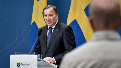 En man i blå kostym står bakom ett podium med Sveriges flagga i bakgrunden. Snett framför honom står en person med ryggen mot kameran i vit skjorta.