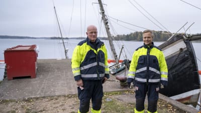 Jörgen Engroos och Fredrik Kevin står på kajen bredvid en stor trålare som ligger halvvägs under vattenytan.