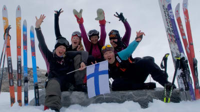 Fem kvinnor iklädda tjocka vinterkläder. Sitter och hurrar och vifta med Finlands flagga. De befinner sig på Grönland, i ett snölandskap.