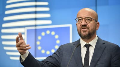 Charles Michel håller tal med handen utsträckt framför en blå bakgrund där EU:s flagga syns. Michel har runda glasögon, skägg och är skallig.