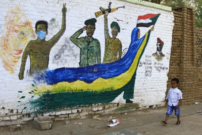 En väggmålning av en några soldater och en kvinna som är klädd i en mantel i blått och gult med Sudans flagga i handen. Förbi väggmålningen går ett barn i blå byxor och vit skjorta.