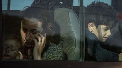 Flyktingar i en buss. Genom bussfönstret syns en kvinna med ett litet barn i famnen. Hon håller en mobiltelefon vid örat och ser trött ut. På raden bakom tittar en pojke på cirka 10 år ut genom fönstret.