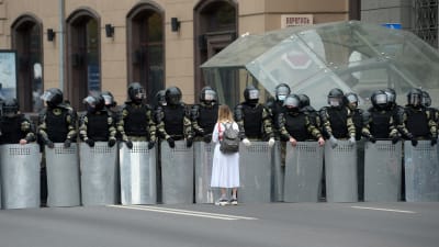 En vitklädd kvinna står framför en rad poliser i full kravallutrustning