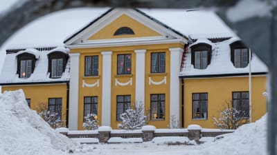 Ett gammalt gult hus täckt i snö. I fönstren lyser julbelysning.