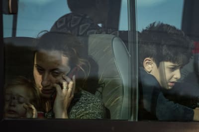Flyktingar i en buss. Genom bussfönstret syns en kvinna med ett litet barn i famnen. Hon håller en mobiltelefon vid örat och ser trött ut. På raden bakom tittar en pojke på cirka 10 år ut genom fönstret.