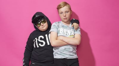 Melvin poserar med solglasögon, keps och huva på huvudet. Oliver står med armarna i kors. Melvin håller sin vänstra arm på Olivers vänstra axel.