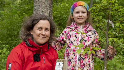 En kvinna i röd rock står med sin sex år gamla dotter i en träddunge och tittar in i kameran.