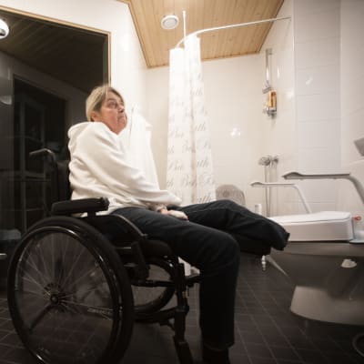 En kvinna sitter i en rullstol, inne i ett badrum. Hon ser uppgiven ut. I bakgrunden syns en dusch, en bastu och en toalettstol.